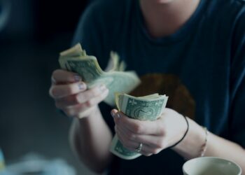 A person wearing a dark shirt is counting a stack of U.S. one-dollar bills, with a coffee cup visible in the foreground. Their face is out of focus and not fully visible. | MONEY6X