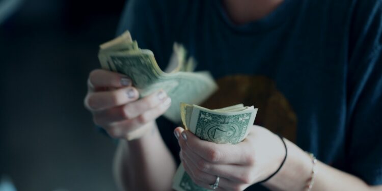 A person wearing a dark shirt is counting a stack of U.S. one-dollar bills, with a coffee cup visible in the foreground. Their face is out of focus and not fully visible. | MONEY6X
