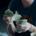 A person wearing a dark shirt is counting a stack of U.S. one-dollar bills, with a coffee cup visible in the foreground. Their face is out of focus and not fully visible. | MONEY6X