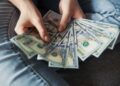 A person wearing jeans holds and fans out several $100 US dollar bills, showing a spread of cash in their hands. | MONEY6X