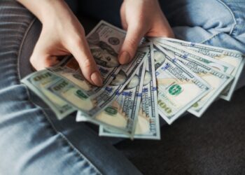 A person wearing jeans holds and fans out several $100 US dollar bills, showing a spread of cash in their hands. | MONEY6X