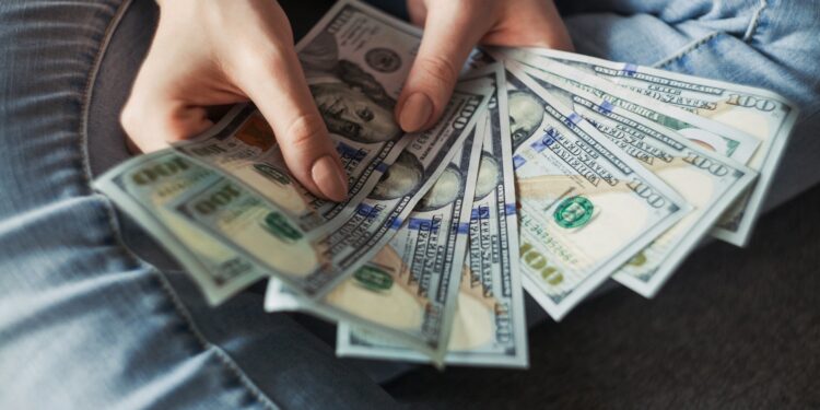 A person wearing jeans holds and fans out several $100 US dollar bills, showing a spread of cash in their hands. | MONEY6X