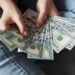 A person wearing jeans holds and fans out several $100 US dollar bills, showing a spread of cash in their hands. | MONEY6X