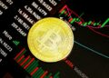 A gold Bitcoin coin rests on a screen displaying financial charts and graphs with various numbers and colored candlestick patterns. | MONEY6X