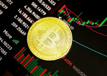 A gold Bitcoin coin rests on a screen displaying financial charts and graphs with various numbers and colored candlestick patterns. | MONEY6X