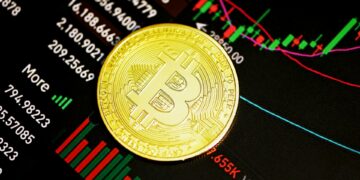 A gold Bitcoin coin rests on a screen displaying financial charts and graphs with various numbers and colored candlestick patterns. | MONEY6X