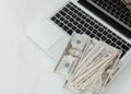 A pile of U.S. dollar bills spread across the keyboard of an open silver laptop on a white surface. | MONEY6X