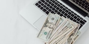 A pile of U.S. dollar bills spread across the keyboard of an open silver laptop on a white surface. | MONEY6X
