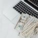 A pile of U.S. dollar bills spread across the keyboard of an open silver laptop on a white surface. | MONEY6X