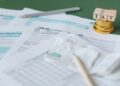 Tax forms, a pen, receipts, and gold coins stacked with wooden blocks spelling “TAX” on top, arranged on a green surface, suggesting tax preparation or financial paperwork. | MONEY6X