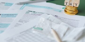 Tax forms, a pen, receipts, and gold coins stacked with wooden blocks spelling “TAX” on top, arranged on a green surface, suggesting tax preparation or financial paperwork. | MONEY6X