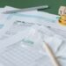 Tax forms, a pen, receipts, and gold coins stacked with wooden blocks spelling “TAX” on top, arranged on a green surface, suggesting tax preparation or financial paperwork. | MONEY6X