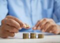A person in a blue shirt stacks coins into neat piles on a white table, focusing on organizing and counting the money. | MONEY6X