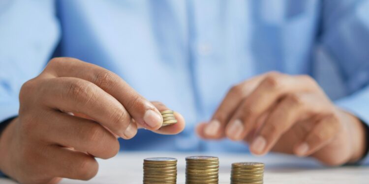 A person in a blue shirt stacks coins into neat piles on a white table, focusing on organizing and counting the money. | MONEY6X