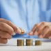 A person in a blue shirt stacks coins into neat piles on a white table, focusing on organizing and counting the money. | MONEY6X