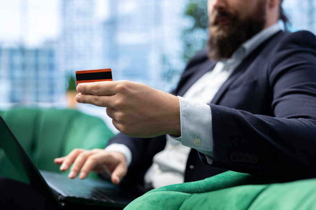 A man in a suit sits on a green chair, holding a credit card in one hand and using a laptop with the other. The background is blurred, showing office windows. | MONEY6X