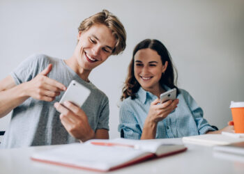 A young man and woman sit at a table looking at a smartphone together, smiling. The man gestures toward the screen while the woman holds the phone. There is an open notebook and an orange coffee cup on the white table in front of them. | MONEY6X