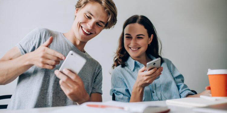 A young man and woman sit at a table looking at a smartphone together, smiling. The man gestures toward the screen while the woman holds the phone. There is an open notebook and an orange coffee cup on the white table in front of them. | MONEY6X