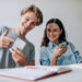 A young man and woman sit at a table looking at a smartphone together, smiling. The man gestures toward the screen while the woman holds the phone. There is an open notebook and an orange coffee cup on the white table in front of them. | MONEY6X