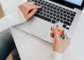 Person in a white sweater holding a credit card and typing on a laptop keyboard, possibly shopping or making an online payment. | MONEY6X