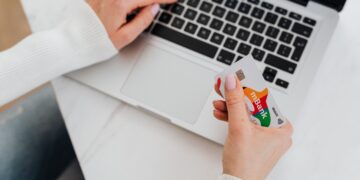 Person in a white sweater holding a credit card and typing on a laptop keyboard, possibly shopping or making an online payment. | MONEY6X