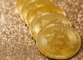 Five gold Bitcoin coins placed in a line on a shimmering, glittery golden surface. | MONEY6X