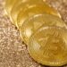 Five gold Bitcoin coins placed in a line on a shimmering, glittery golden surface. | MONEY6X