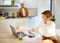 A woman smiles while talking on the phone, sitting at a table with a laptop, papers, calculator, and a green drink—possibly discussing credit card balances. A man stands in the background in a bright, modern kitchen. | MONEY6X