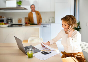 A woman smiles while talking on the phone, sitting at a table with a laptop, papers, calculator, and a green drink—possibly discussing credit card balances. A man stands in the background in a bright, modern kitchen. | MONEY6X