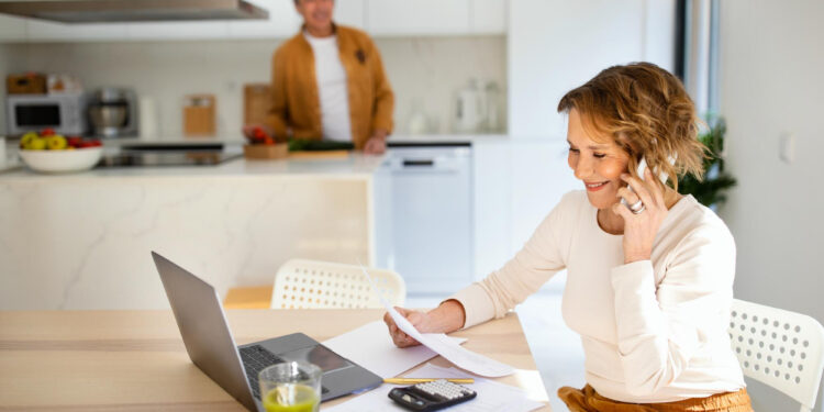 A woman smiles while talking on the phone, sitting at a table with a laptop, papers, calculator, and a green drink—possibly discussing credit card balances. A man stands in the background in a bright, modern kitchen. | MONEY6X
