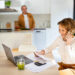 A woman smiles while talking on the phone, sitting at a table with a laptop, papers, calculator, and a green drink—possibly discussing credit card balances. A man stands in the background in a bright, modern kitchen. | MONEY6X